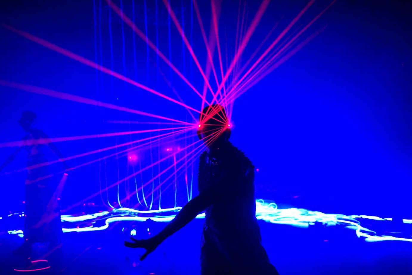 Blue light background a central figure in silouhette red beams of light rise from the figures head and eyes surrealistically